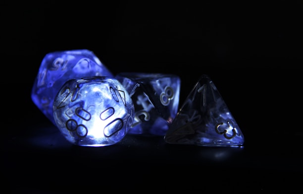Illuminated polyhedral dice glowing with a bluish hue against a dark background. The light creates a futuristic and mystical atmosphere highlighting the numbers on the intricately shaped dice.