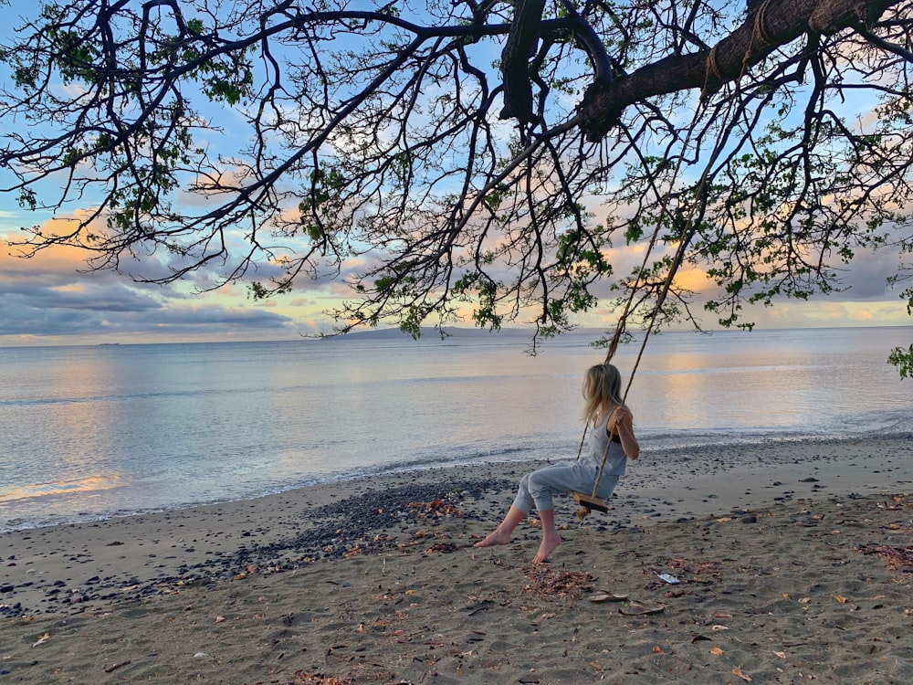 girl wearing blue romper pants on swing hanging on tree besides seashore