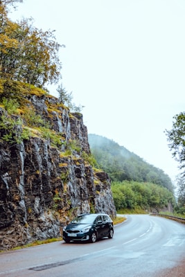 A car is parked on a winding road next to a steep rock face and lush greenery. The road curves gently into the distance, bordered by a dense forest with mist hovering around the trees. The scene is serene and peaceful, capturing the essence of a road trip through nature.