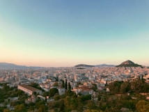 The image captures a panoramic view of a cityscape during sunset, with closely packed buildings and greenery. Notable are two hills rising in the distance, creating a dynamic horizon. The sky displays a gradient of warm colors transitioning to cooler hues at the top.