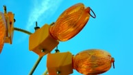 Several orange construction lights attached to square yellow bases are mounted on a white pole against a bright blue sky.