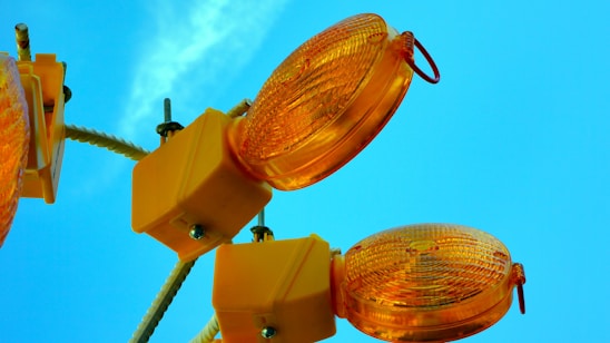 Several orange construction lights attached to square yellow bases are mounted on a white pole against a bright blue sky.