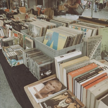 A market stall filled with an assortment of vintage books and vinyl records. Several stacks and bins hold items, with some records displaying visible covers. Customers can be seen in the background browsing through the collection.