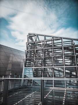 A modern architectural structure composed of a geometric grid of glass panels reflecting the ambient sky. The design features overlapping metal and glass elements situated in an urban environment. The image captures a sense of modernity and innovation.