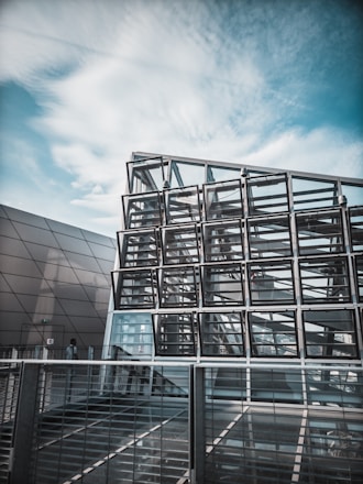 A modern architectural structure composed of a geometric grid of glass panels reflecting the ambient sky. The design features overlapping metal and glass elements situated in an urban environment. The image captures a sense of modernity and innovation.