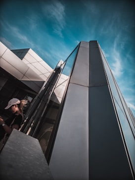 A modern architectural structure with sleek, angular lines and reflective glass panels stretches into a blue, partly cloudy sky. A person wearing a white cap and dark shirt is seen in the foreground, possibly interacting with a mobile device.