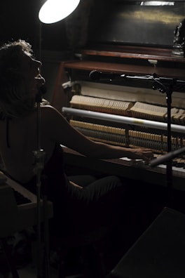 A musician is playing a piano with exposed strings, accompanied by a microphone on a stand. The scene is dimly lit with a bright overhead lamp casting light on the performer. The overall ambiance suggests an intimate musical setting.