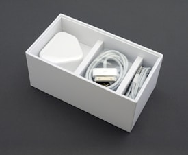 A neatly arranged white box containing a power adapter, a coiled charging cable, and an audio cable. The box is sectioned into compartments, neatly organizing the electronic accessories.