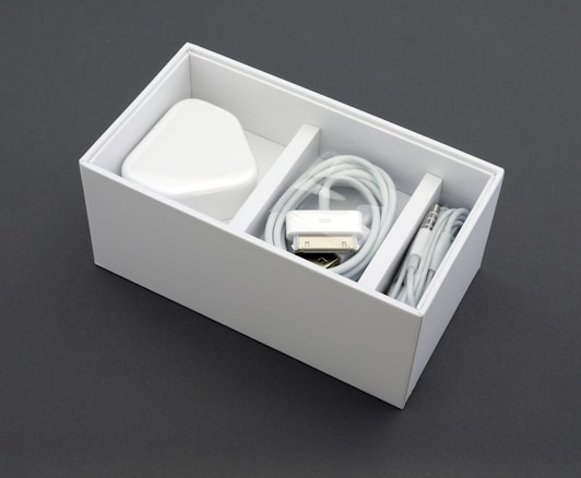 A neatly arranged white box containing a power adapter, a coiled charging cable, and an audio cable. The box is sectioned into compartments, neatly organizing the electronic accessories.