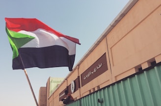 A large flag with red, white, black, and green colors is being held up against a clear sky. The background features a tan building with Arabic writing and some green structural elements.