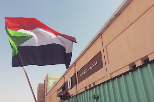 A large flag with red, white, black, and green colors is being held up against a clear sky. The background features a tan building with Arabic writing and some green structural elements.