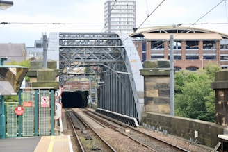 A railway track leads towards an ornate metal bridge with intricate framework. There are signs and a barrier at the station platform, and overhead power lines stretch along the track. In the background, there are trees and various buildings, including a large brick structure.