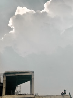 A large, dramatic cloud dominates the sky above a partially constructed building. The structure has an exposed concrete frame, and several people are visible on the right side, wearing construction gear. The atmosphere is calm, with soft lighting creating a tranquil scene.