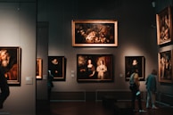 A dimly lit art gallery with several framed paintings hanging on dark walls. A few visitors are observing the artwork, with one person taking a photo using a smartphone. The paintings depict various classical portraiture styles.