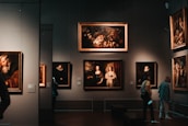 A dimly lit art gallery with several framed paintings hanging on dark walls. A few visitors are observing the artwork, with one person taking a photo using a smartphone. The paintings depict various classical portraiture styles.