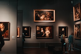 A dimly lit art gallery with several framed paintings hanging on dark walls. A few visitors are observing the artwork, with one person taking a photo using a smartphone. The paintings depict various classical portraiture styles.