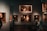 A dimly lit art gallery with several framed paintings hanging on dark walls. A few visitors are observing the artwork, with one person taking a photo using a smartphone. The paintings depict various classical portraiture styles.