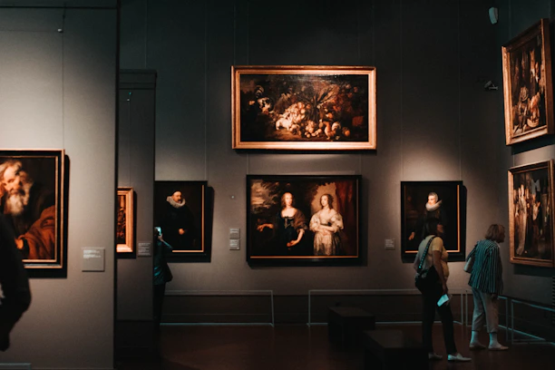 A dimly lit art gallery with several framed paintings hanging on dark walls. A few visitors are observing the artwork, with one person taking a photo using a smartphone. The paintings depict various classical portraiture styles.
