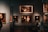 A dimly lit art gallery with several framed paintings hanging on dark walls. A few visitors are observing the artwork, with one person taking a photo using a smartphone. The paintings depict various classical portraiture styles.
