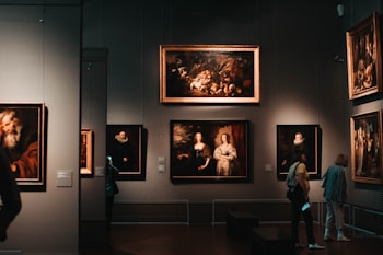 A dimly lit art gallery with several framed paintings hanging on dark walls. A few visitors are observing the artwork, with one person taking a photo using a smartphone. The paintings depict various classical portraiture styles.