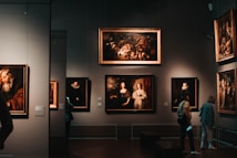 A dimly lit art gallery with several framed paintings hanging on dark walls. A few visitors are observing the artwork, with one person taking a photo using a smartphone. The paintings depict various classical portraiture styles.