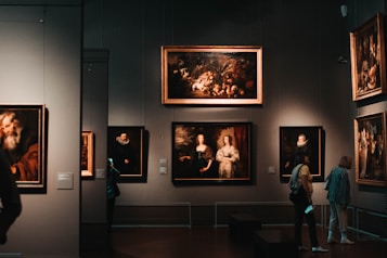 A dimly lit art gallery with several framed paintings hanging on dark walls. A few visitors are observing the artwork, with one person taking a photo using a smartphone. The paintings depict various classical portraiture styles.