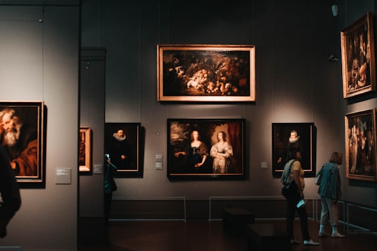 A dimly lit art gallery with several framed paintings hanging on dark walls. A few visitors are observing the artwork, with one person taking a photo using a smartphone. The paintings depict various classical portraiture styles.