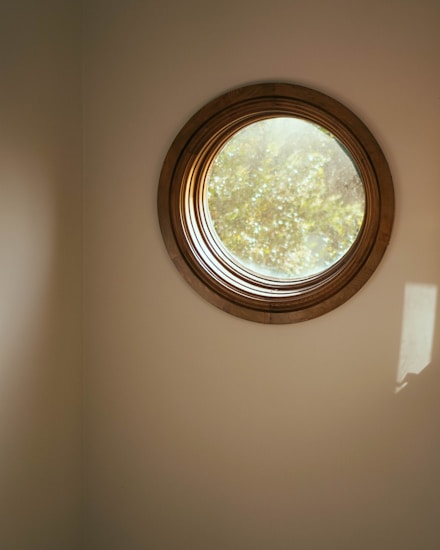 A circular window with a wooden frame emits soft natural light into an interior space. The light outside appears dappled, suggesting the presence of leaves or branches. The walls are a muted beige color, enhancing the warmth of the light.