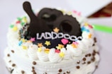 A decorated cake with white icing and colorful star-shaped sprinkles. Chocolate decorations and lettering are on top, with chocolate chips around the sides.