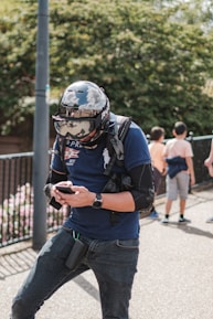 A person wearing a large helmet and protective gear stands outdoors, interacting with a smartphone. The helmet has reflective properties, showing parts of the surroundings. The individual is dressed casually in jeans and a t-shirt and wears a backpack. In the background, a few other people, including children, are visible near green foliage and a fence.