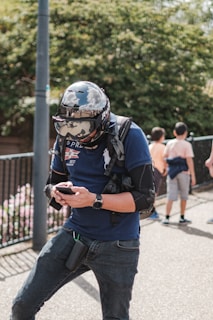 A person wearing a large helmet and protective gear stands outdoors, interacting with a smartphone. The helmet has reflective properties, showing parts of the surroundings. The individual is dressed casually in jeans and a t-shirt and wears a backpack. In the background, a few other people, including children, are visible near green foliage and a fence.