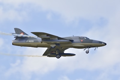 A military jet in mid-flight against a backdrop of cloudy sky. The aircraft is painted in a muted camouflage color with specific markings, including a red cross and numbers on the fuselage. Smoke trails from the wing and it appears to be an older model jet due to its design features.