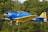 A vintage blue and yellow aircraft is flying against a backdrop of lush green trees. The airplane features insignia and has an open cockpit with a pilot wearing sunglasses.