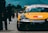 A yellow race car with numerous sponsorship stickers is prominently displayed on a race track. People can be seen walking and standing in the background, likely members of a racing team or spectators. The backdrop includes trees and rolling hills under a blue sky.