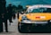A yellow race car with numerous sponsorship stickers is prominently displayed on a race track. People can be seen walking and standing in the background, likely members of a racing team or spectators. The backdrop includes trees and rolling hills under a blue sky.