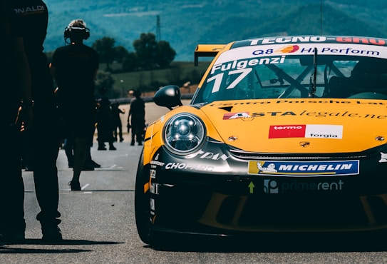 A yellow race car with numerous sponsorship stickers is prominently displayed on a race track. People can be seen walking and standing in the background, likely members of a racing team or spectators. The backdrop includes trees and rolling hills under a blue sky.