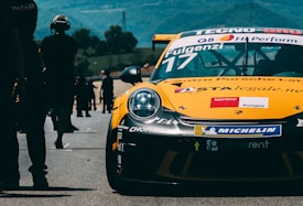 A yellow race car with numerous sponsorship stickers is prominently displayed on a race track. People can be seen walking and standing in the background, likely members of a racing team or spectators. The backdrop includes trees and rolling hills under a blue sky.