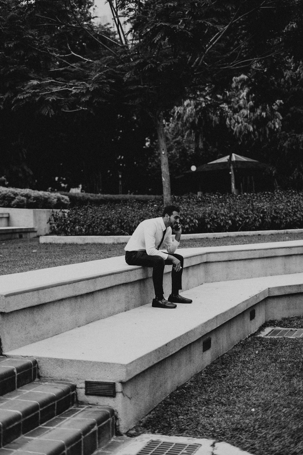 grayscale photography of man using phone while sitting on concrete pavement