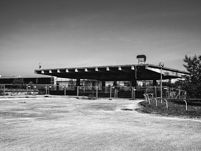 A black and white photograph of an empty, abandoned lot featuring a large, flat-roofed structure. The area is surrounded by a metal fence, and the ground is cracked with some overgrown grass. The structure appears industrial or commercial, with a simple, utilitarian design.