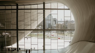 A view from inside a modern architectural building with sweeping curves, looking out through large glass windows towards an urban landscape. The cityscape features tall buildings, open green spaces, and distant structures. Several people are walking or standing near the windows, adding a sense of scale to the massive interior space.