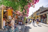 A vibrant street scene in a historical town with people walking and cycling. The image features a quaint marketplace with colorful clothing, bags, and various goods displayed outside shops. Bougainvillea hangs abundantly from above, adding a burst of pink to the setting. The buildings have traditional architectural elements, painted in warm earth tones.