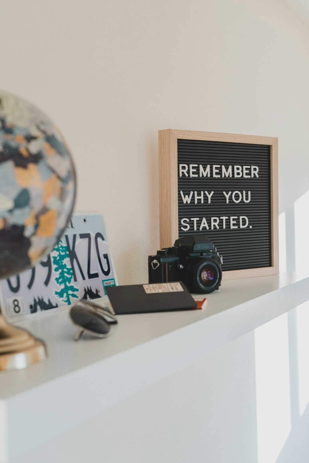 Photo of objects on a mantle, including Oregon license plate, globe, passport, black camera, and a black frame with white lettering, "Remember why you started."