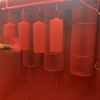 Several cylindrical objects are suspended on hooks in an enclosed space, while a spray tool applies a reddish powder coating to them. The environment is saturated with the same red hue, creating a uniform, monochromatic appearance.