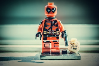 A toy figurine resembling a superhero in a red suit, holding a black accessory, is positioned on a display stand. Next to it, there is a detachable headpiece with an angry expression. The background is softly blurred, highlighting the figurine.