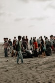 A group of people gathered on a sandy beach, engaged in a cleanup activity. They are surrounded by large black garbage bags filled with collected trash. The sky is overcast, giving the scene a muted background.