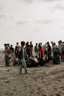 A group of people gathered on a sandy beach, engaged in a cleanup activity. They are surrounded by large black garbage bags filled with collected trash. The sky is overcast, giving the scene a muted background.