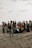A group of people gathered on a sandy beach, engaged in a cleanup activity. They are surrounded by large black garbage bags filled with collected trash. The sky is overcast, giving the scene a muted background.
