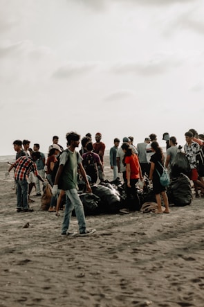 A group of people gathered on a sandy beach, engaged in a cleanup activity. They are surrounded by large black garbage bags filled with collected trash. The sky is overcast, giving the scene a muted background.