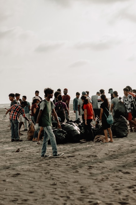 A group of people gathered on a sandy beach, engaged in a cleanup activity. They are surrounded by large black garbage bags filled with collected trash. The sky is overcast, giving the scene a muted background.