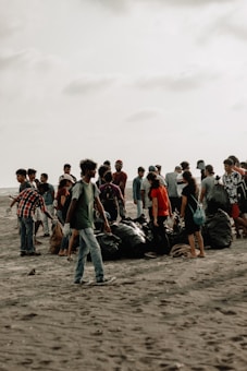 A group of people gathered on a sandy beach, engaged in a cleanup activity. They are surrounded by large black garbage bags filled with collected trash. The sky is overcast, giving the scene a muted background.